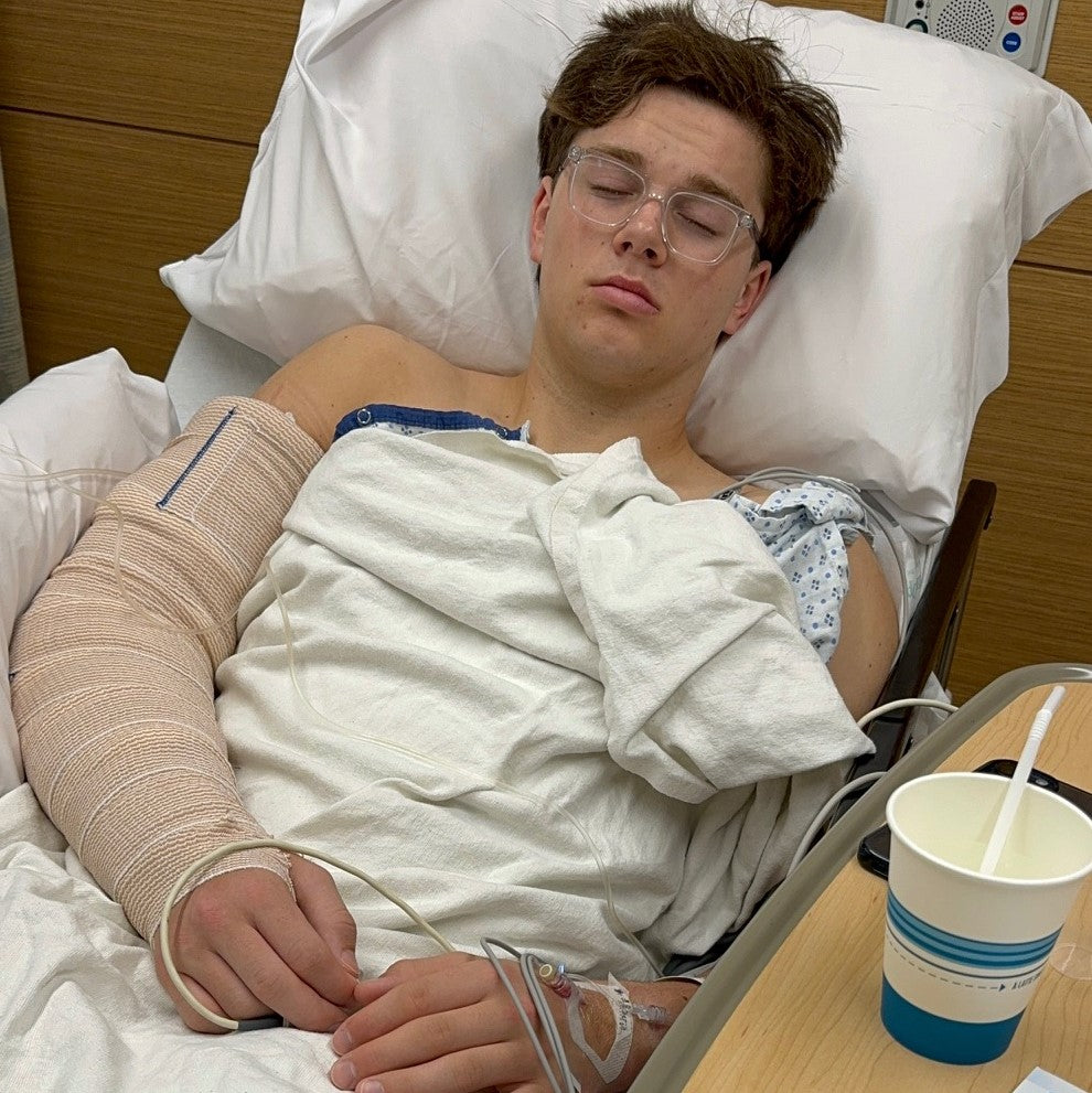 Young pitcher asleep in a hospital bed after Tommy John elbow surgery, his arm wrapped in a post-surgical bandage with an IV line in his hand.