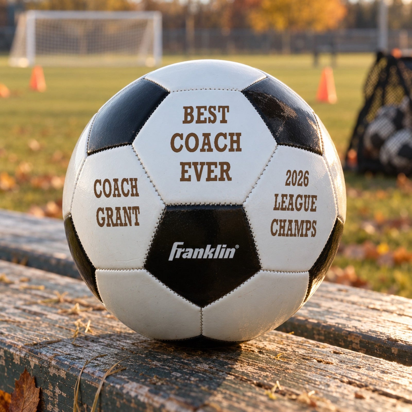 Coach soccer ball engraved with Best Coach Ever, the coach's name and 2026 League Champs, resting on a team bench at the field, a soccer coach gift