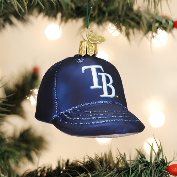 Navy blue baseball cap ornament with white "TB" logo and glitter accent on brim, hanging from evergreen garland with warm white lights