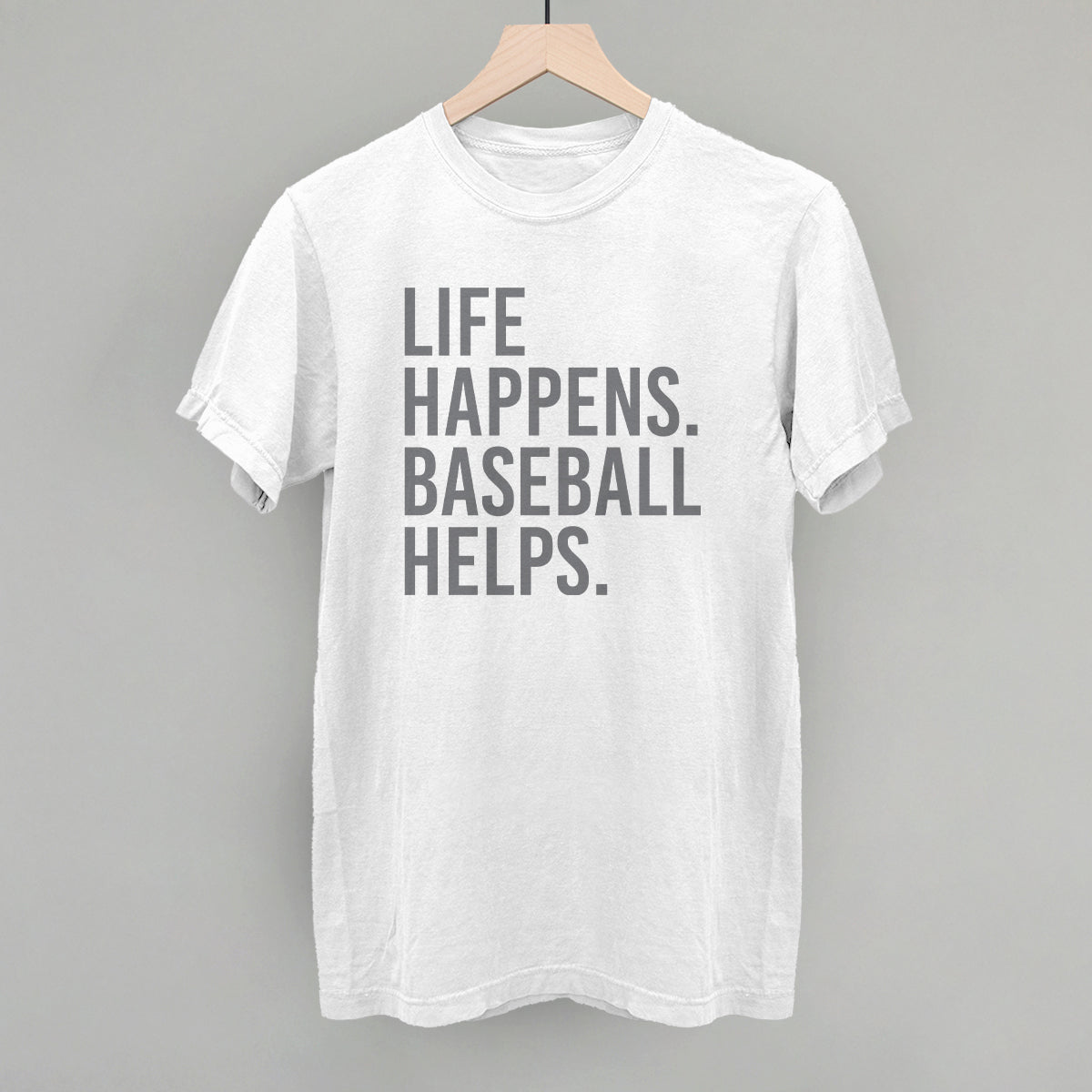 White crewneck t-shirt with gray printed text reading "LIFE HAPPENS. BASEBALL HELPS." displayed on wooden hanger against gray background.