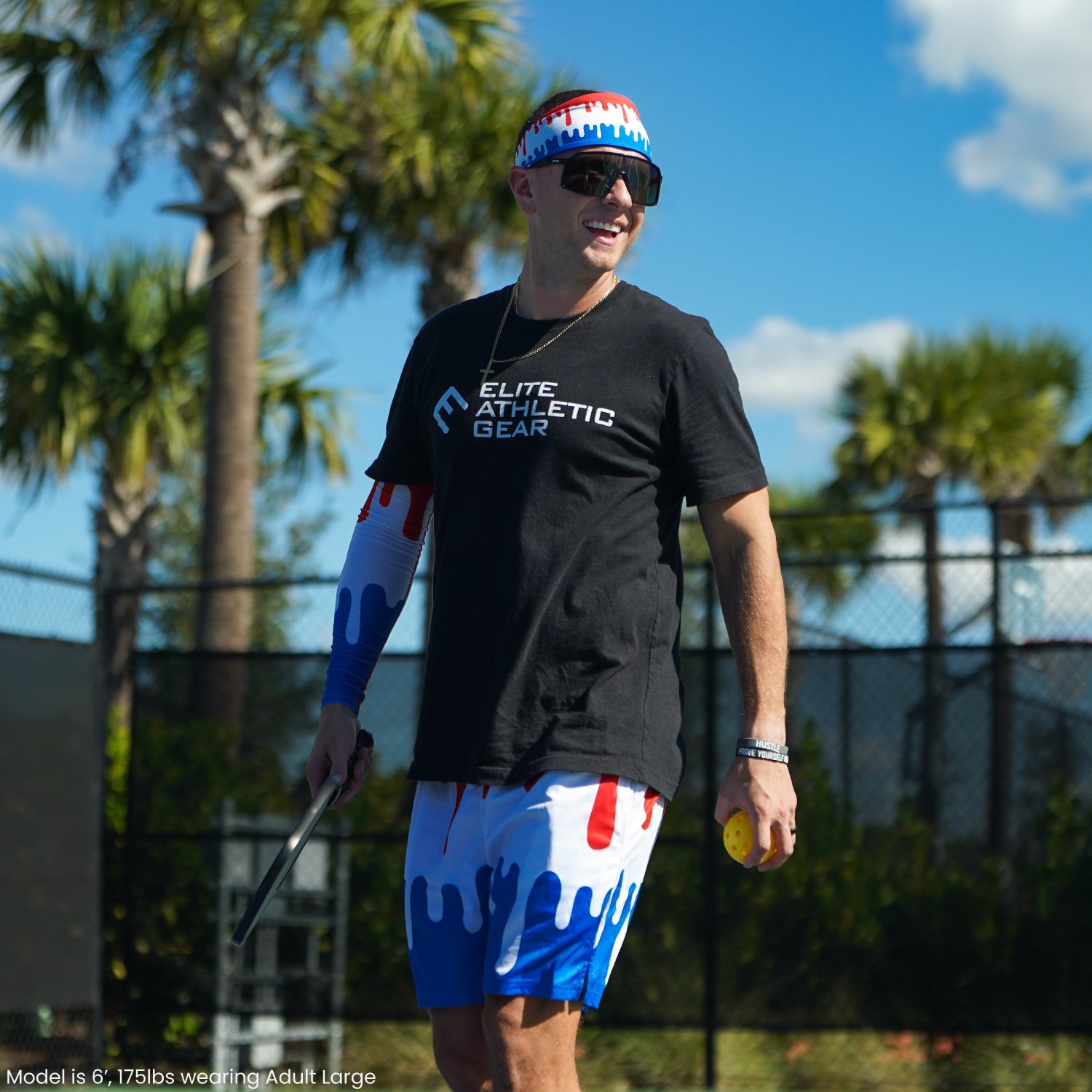 Man wearing blue and white drip-pattern shorts with red accents and matching arm sleeve, standing at baseball field with palm trees in background