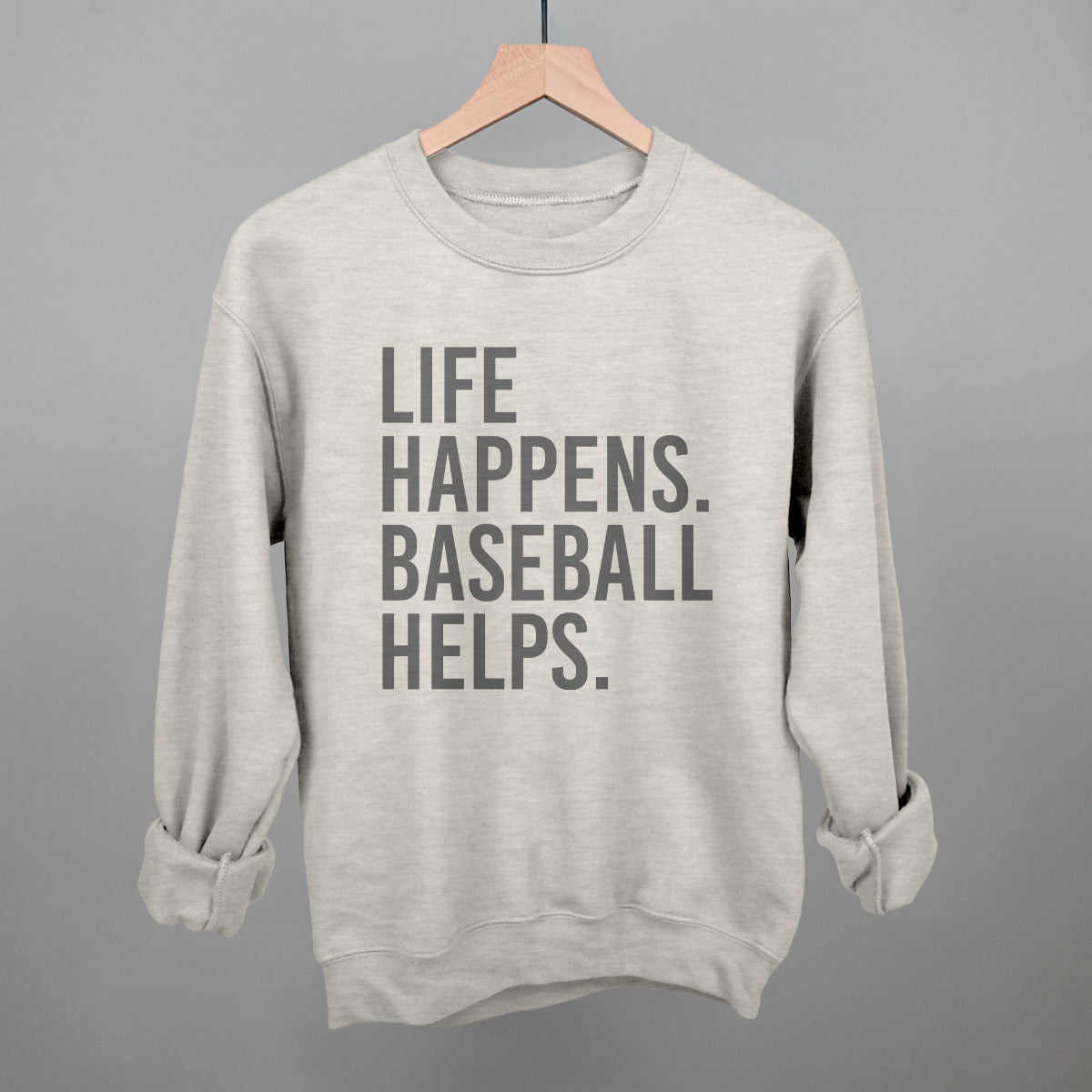 Cream-colored crew sweatshirt with gray printed text reading "LIFE HAPPENS. BASEBALL HELPS." displayed on wooden hanger against neutral background.
