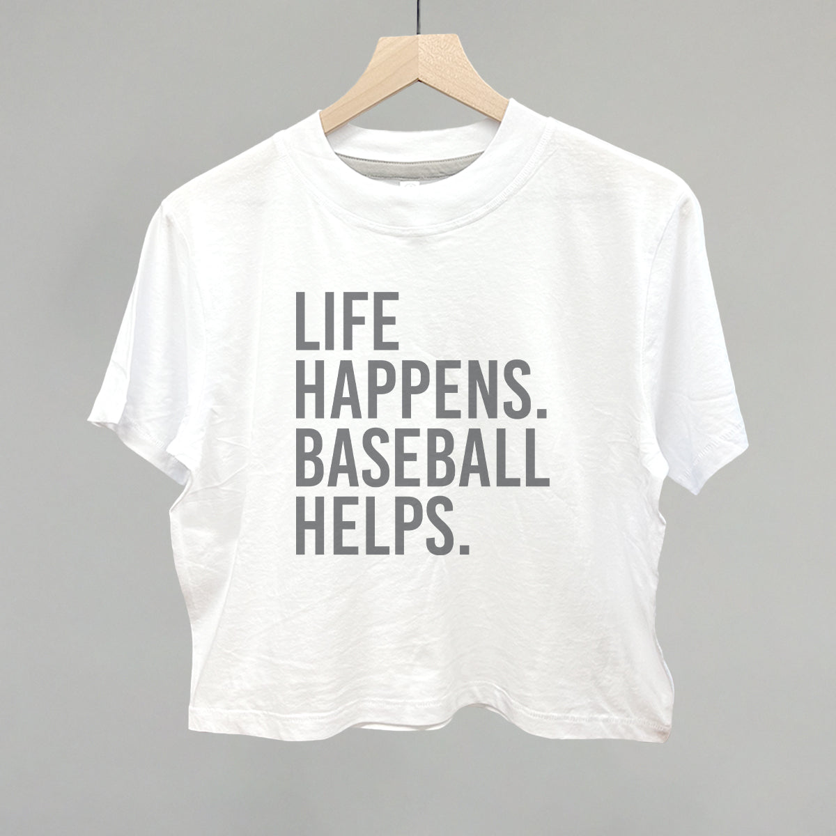 White crewneck tee with gray printed text reading "LIFE HAPPENS. BASEBALL HELPS." displayed on wooden hanger against gray background
