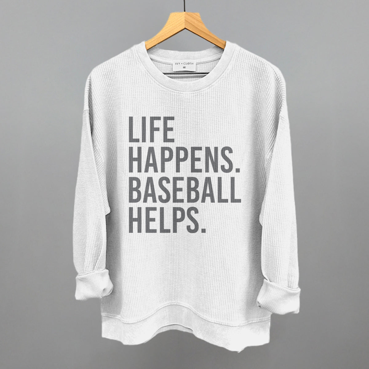 Light gray ribbed crewneck sweatshirt with gray printed text reading "LIFE HAPPENS. BASEBALL HELPS." displayed on wooden hanger against gray background.