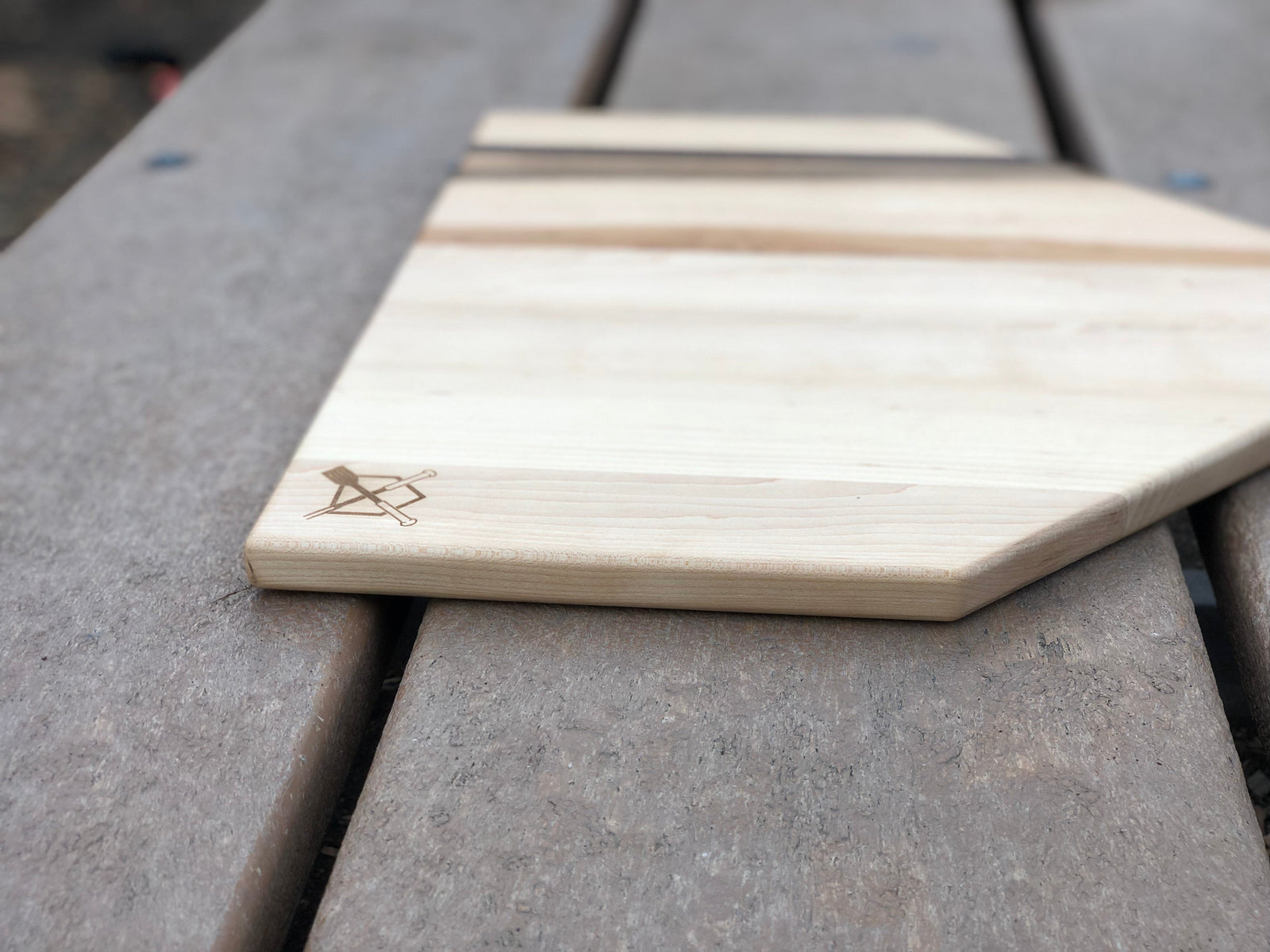 Natural maple home plate cutting board with engraved sample logo in corner, displayed on gray surface alongside stacked boards