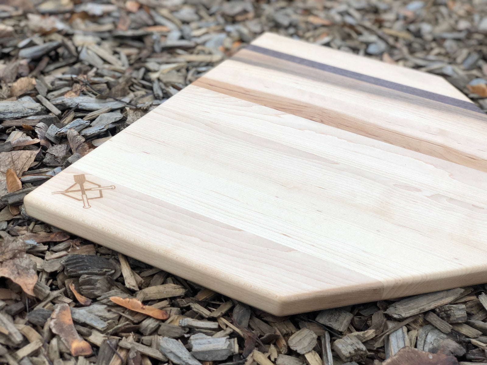 Natural maple home plate cutting board with black walnut accent stripe and sample engraved logo, displayed on wood chips.