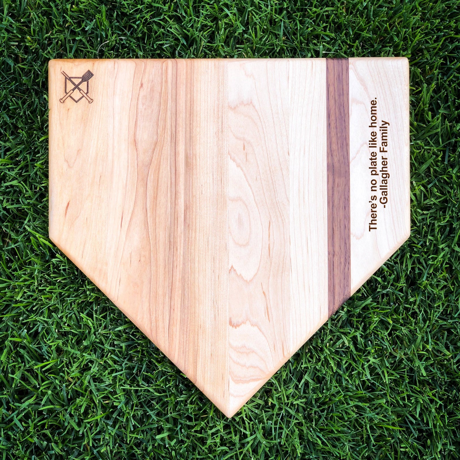 Natural wood home plate cutting board with sample engraving reading "There's no plate like home. -Gallagher Family" along the right edge on grass background.