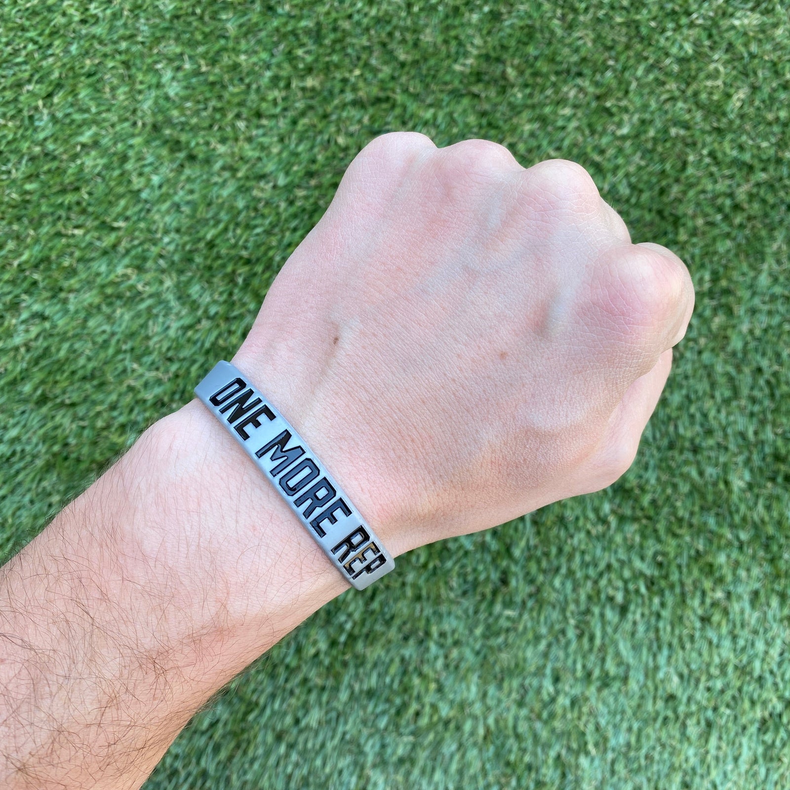 Light blue silicone wristband with debossed black text reading "ONE MORE REP" worn on a person's wrist against green turf background
