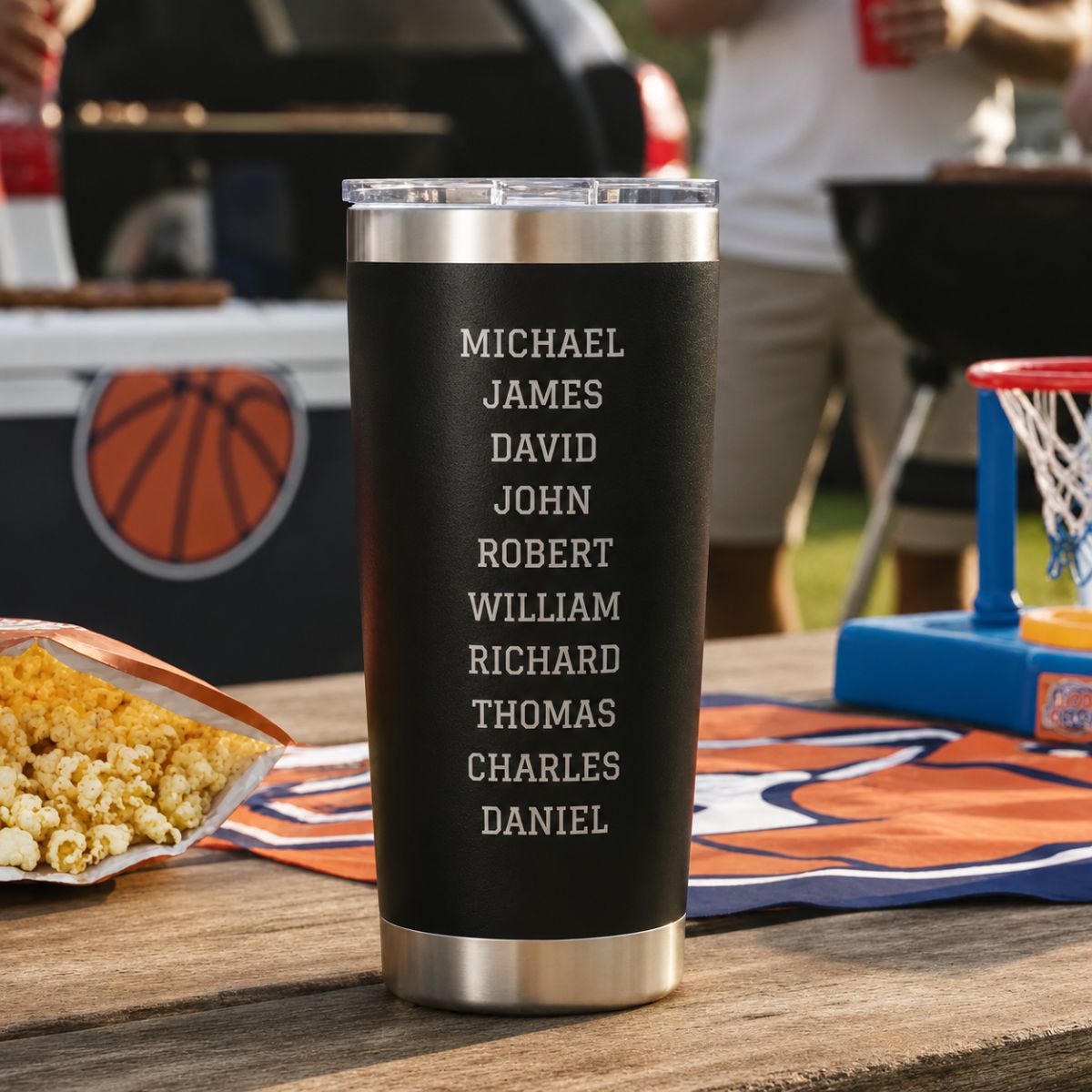 Team roster side of the personalized basketball coach tumbler with ten engraved player names, on a picnic table with popcorn and a basketball at a party