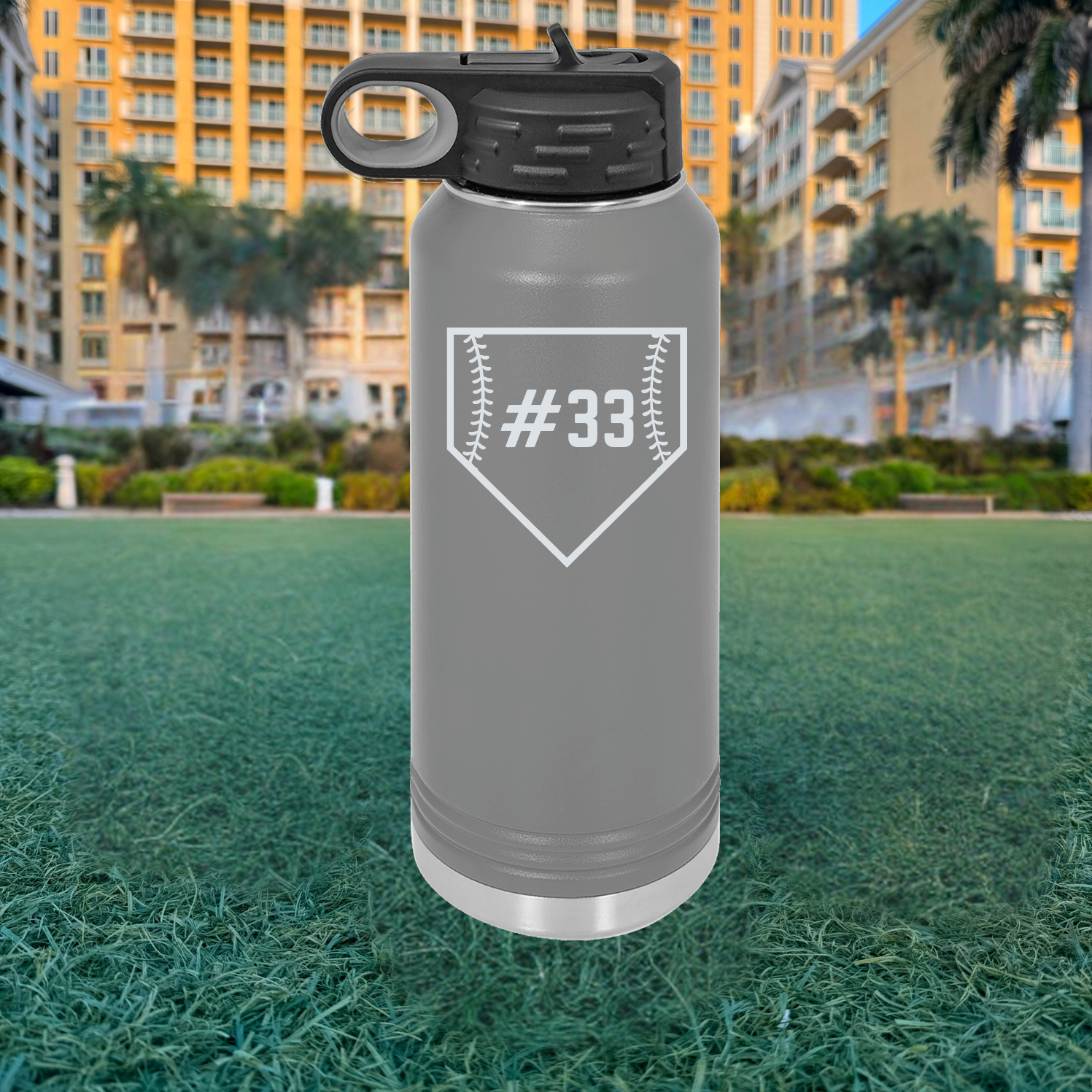 Gray stainless steel water bottle with black flip-top lid, engraved with sample baseball home plate design containing number 33, positioned on grass field with buildings in background.