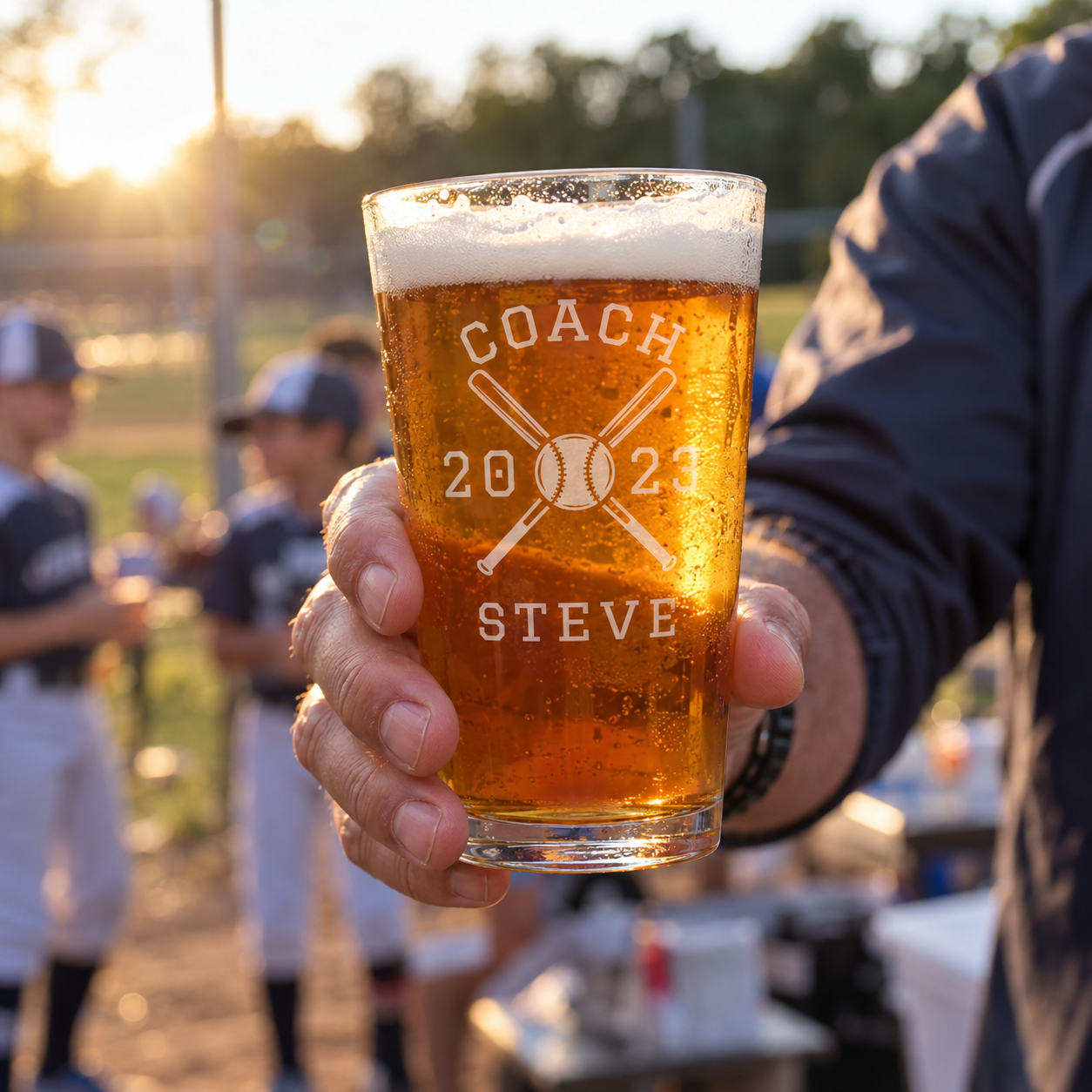 Team Roster Baseball Coach Pint Glass held up at the ballfield after a game, front engraved COACH with crossed bats, 2023 and STEVE, players behind