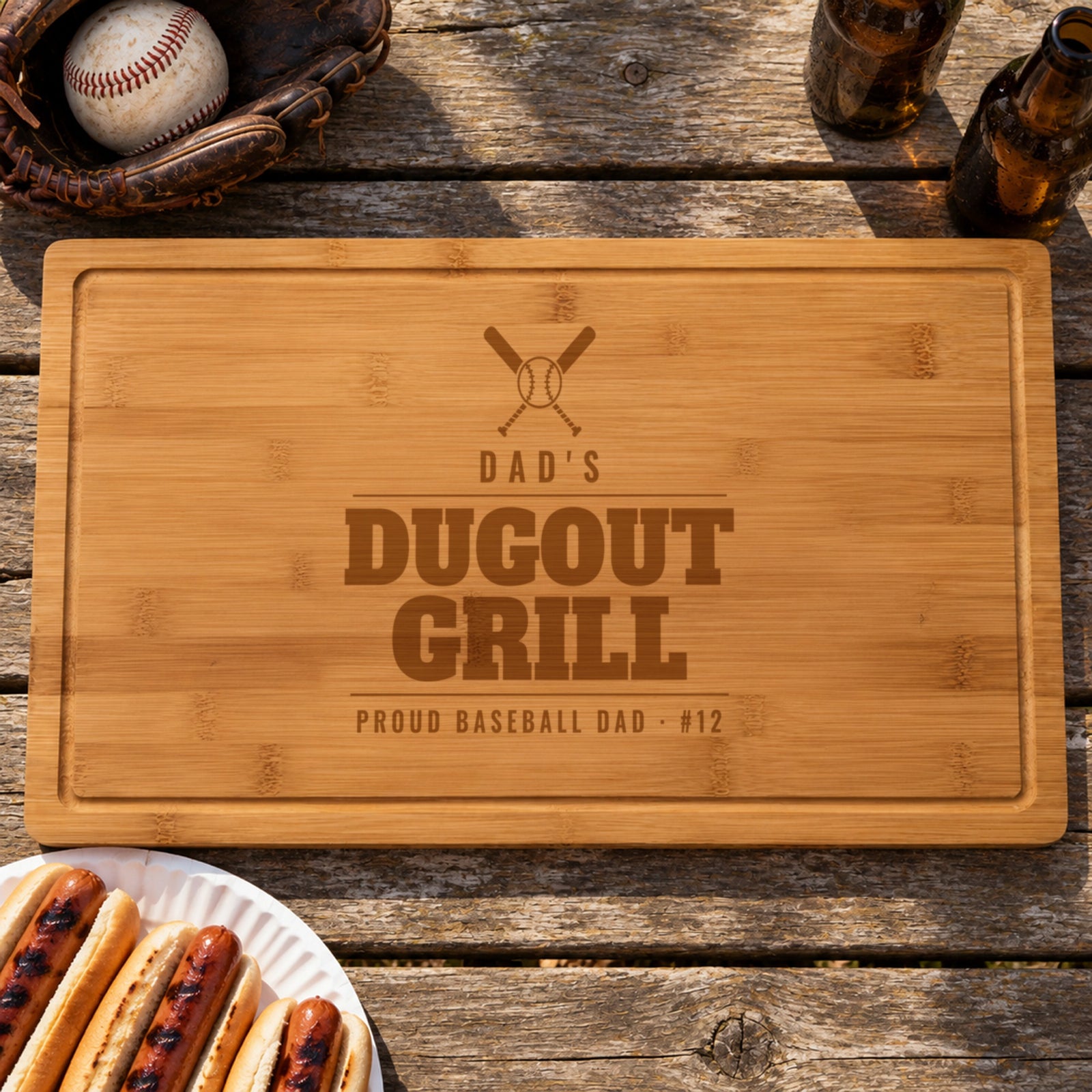 Baseball dad gift: bamboo cutting board engraved with crossed bats, DAD'S DUGOUT GRILL and PROUD BASEBALL DAD #12, beside a ball glove and hot dogs