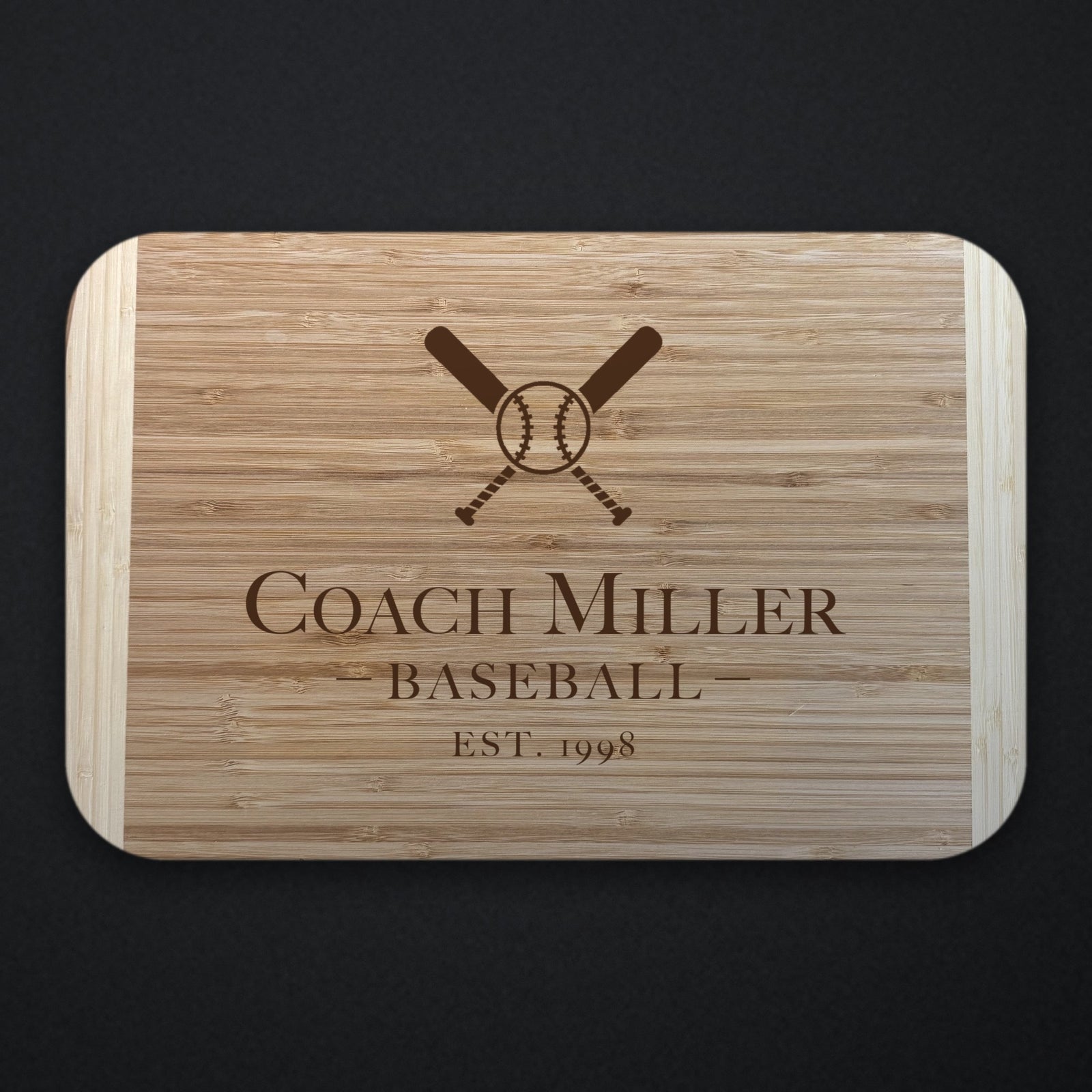 Two tone bamboo baseball coach cutting board engraved with crossed bats and a ball above the name Coach Miller, with BASEBALL and EST. 1998 below, on a dark background.