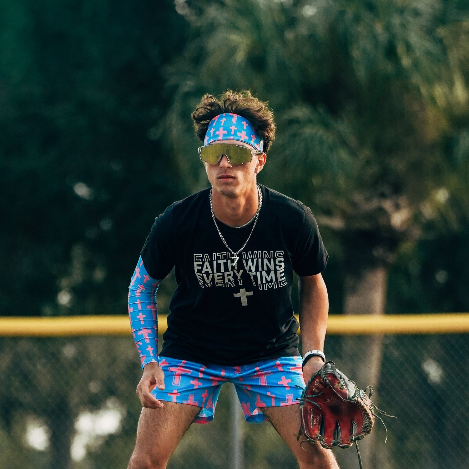 Man wearing Cotton Candy Cross Headband with matching arm sleeve and shorts in blue with pink cross pattern, standing on baseball field with glove