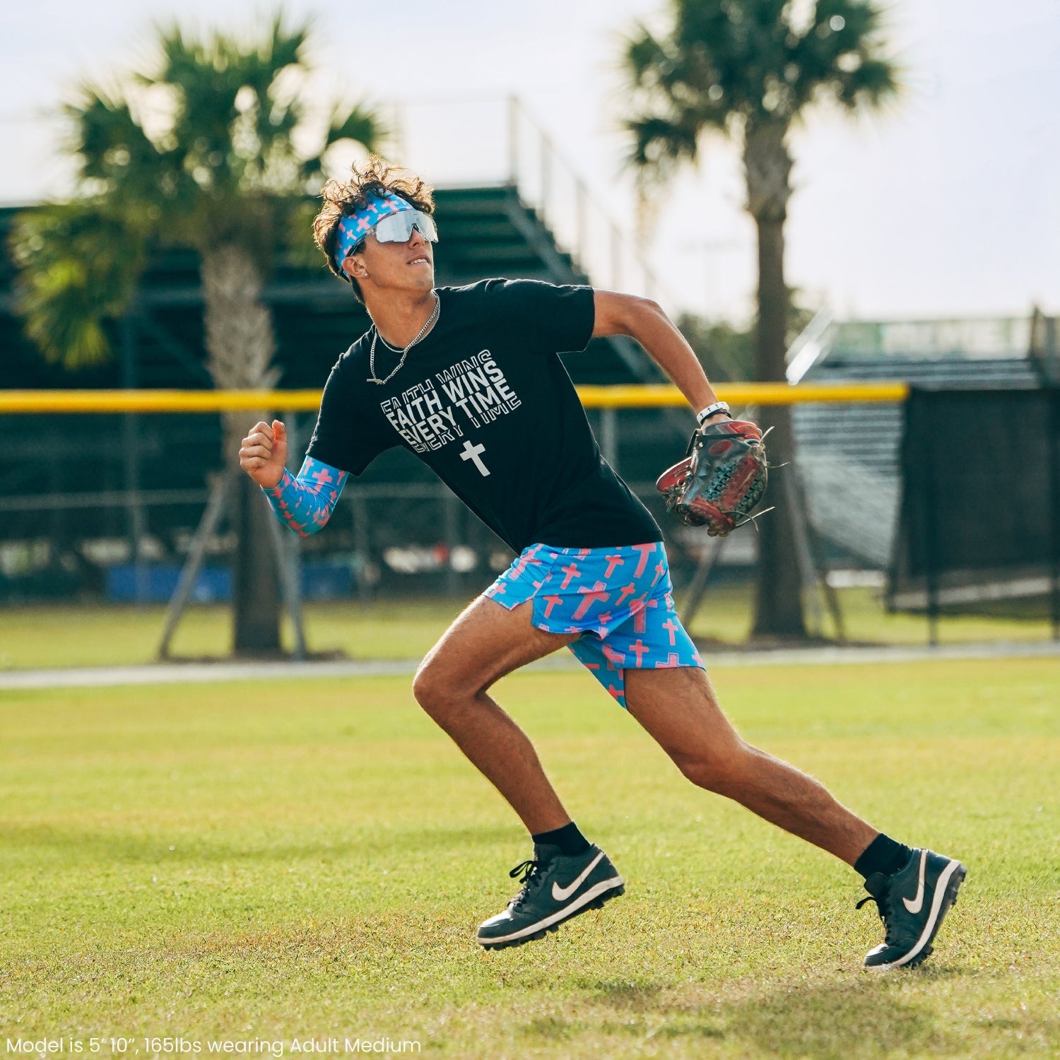 Player wearing Cotton Candy Cross shorts in blue with red cross pattern, paired with black tee and matching headband, fielding a baseball during outdoor practice