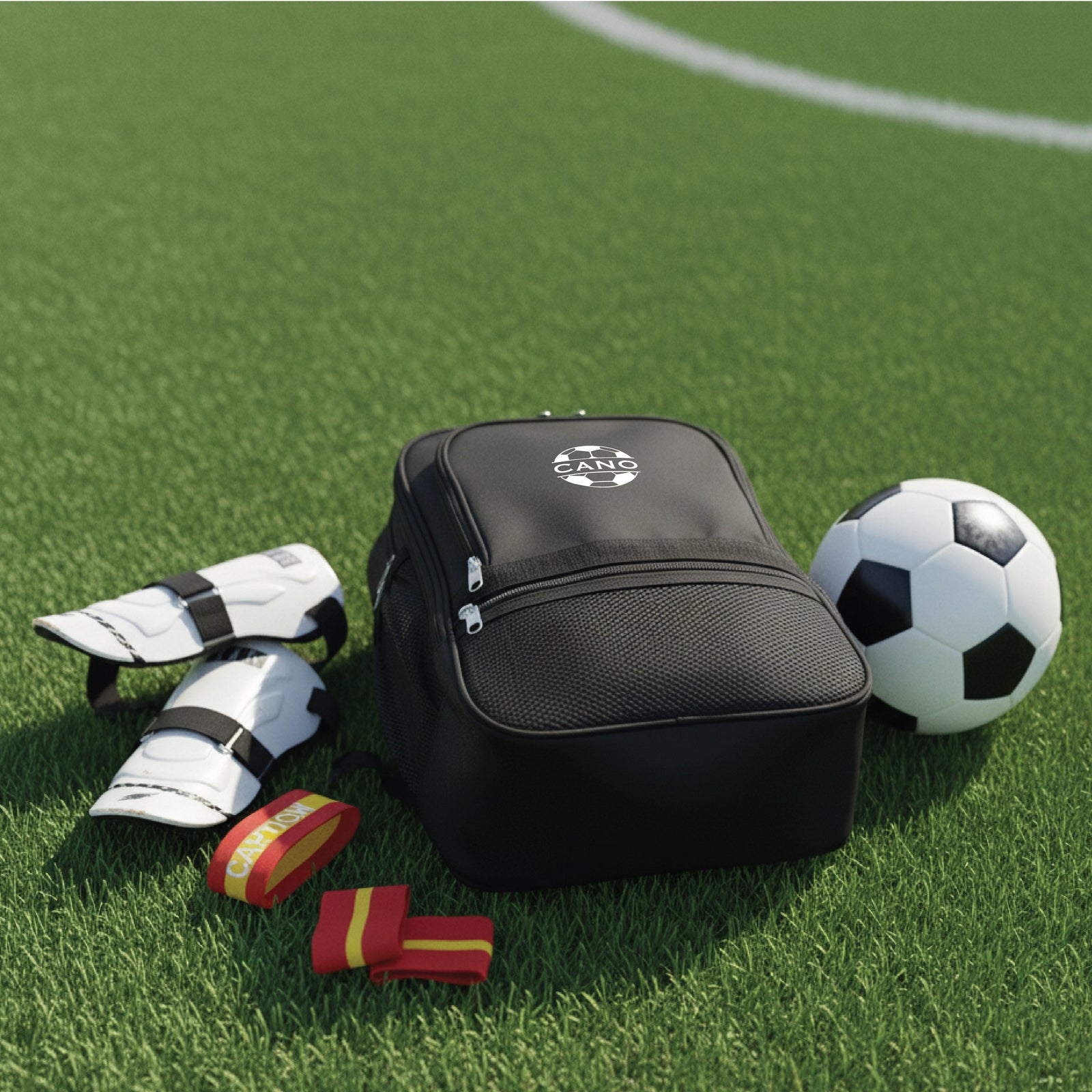 Black soccer equipment bag displayed on artificial turf with shin guards, soccer ball, and red and gold arm sleeves