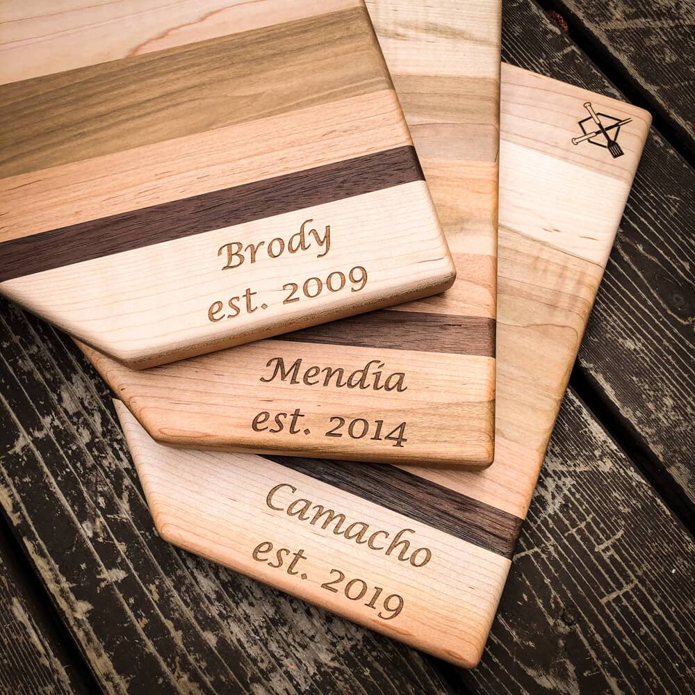 Stack of three home plate shaped cutting boards with sample engraved names and establishment years: "Brody est. 2009," "Mendia est. 2014," and "Camacho est. 2019," showing wood grain variation.