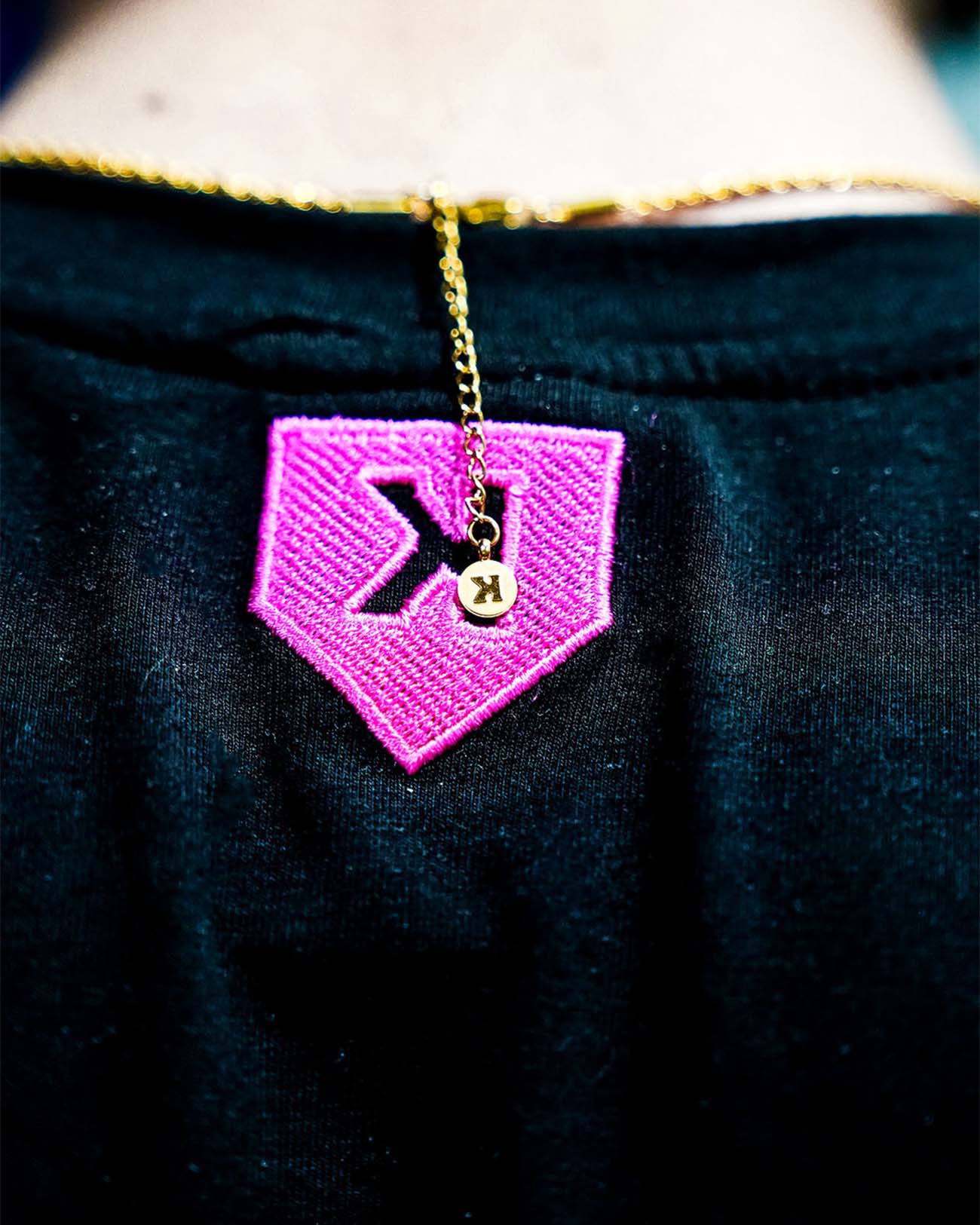 Bright magenta embroidered patch with backwards K design and gold-plated charm pendant hanging from gold rope chain, displayed on dark fabric.