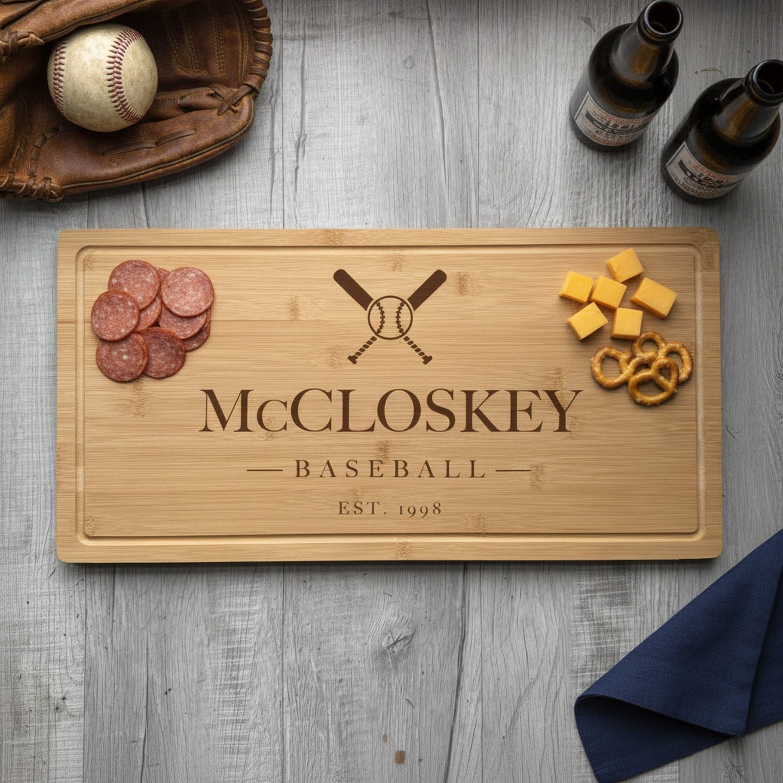 Engraved baseball bamboo board on weathered wood with sliced summer sausage at one end, cheddar and pretzels at the other, beside a glove and two beers.