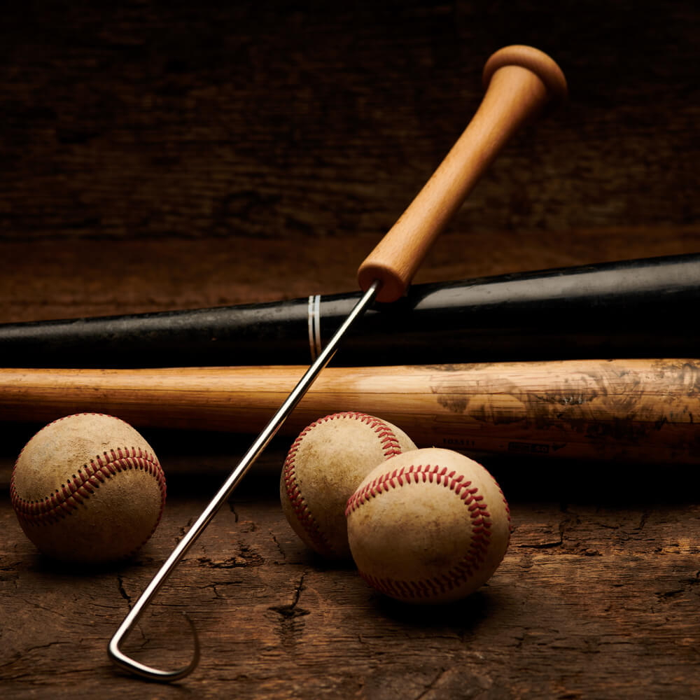 Bat-handle pigtail meat hook leaned across vintage baseball bats and worn baseballs on a dark wood plank backdrop