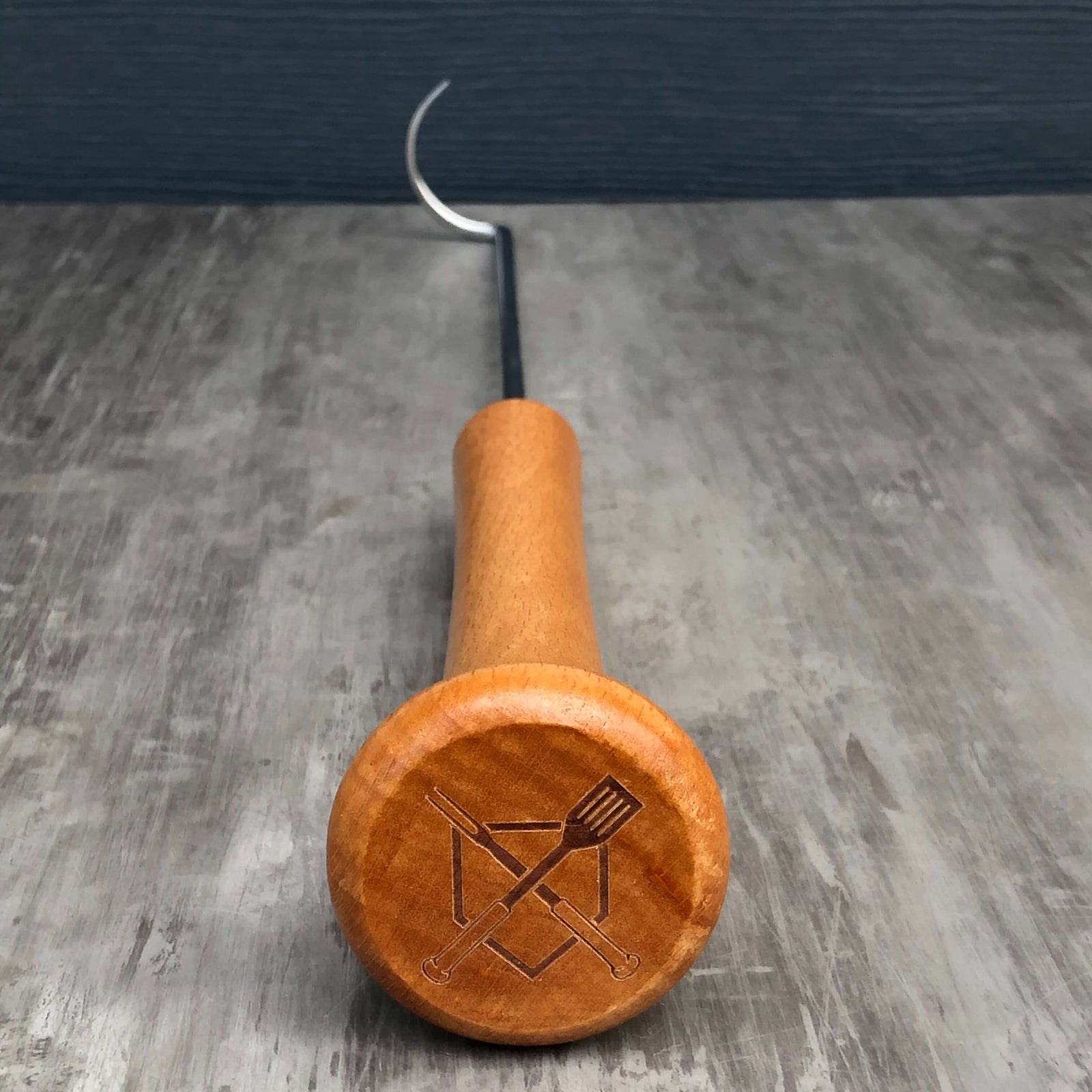 End-grain view of the pigtail's bat-knob handle laser-engraved with the BBS crossed-bat-and-spatula logo on a grey wood-grain surface