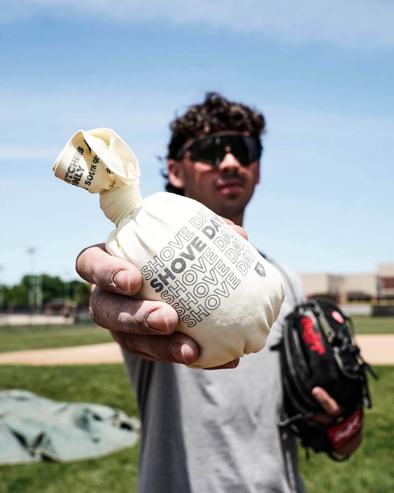 Cream-colored sock-style rosin bag with "SHOVE" text printed repeatedly, held in pitcher's hand at baseball field during daytime.