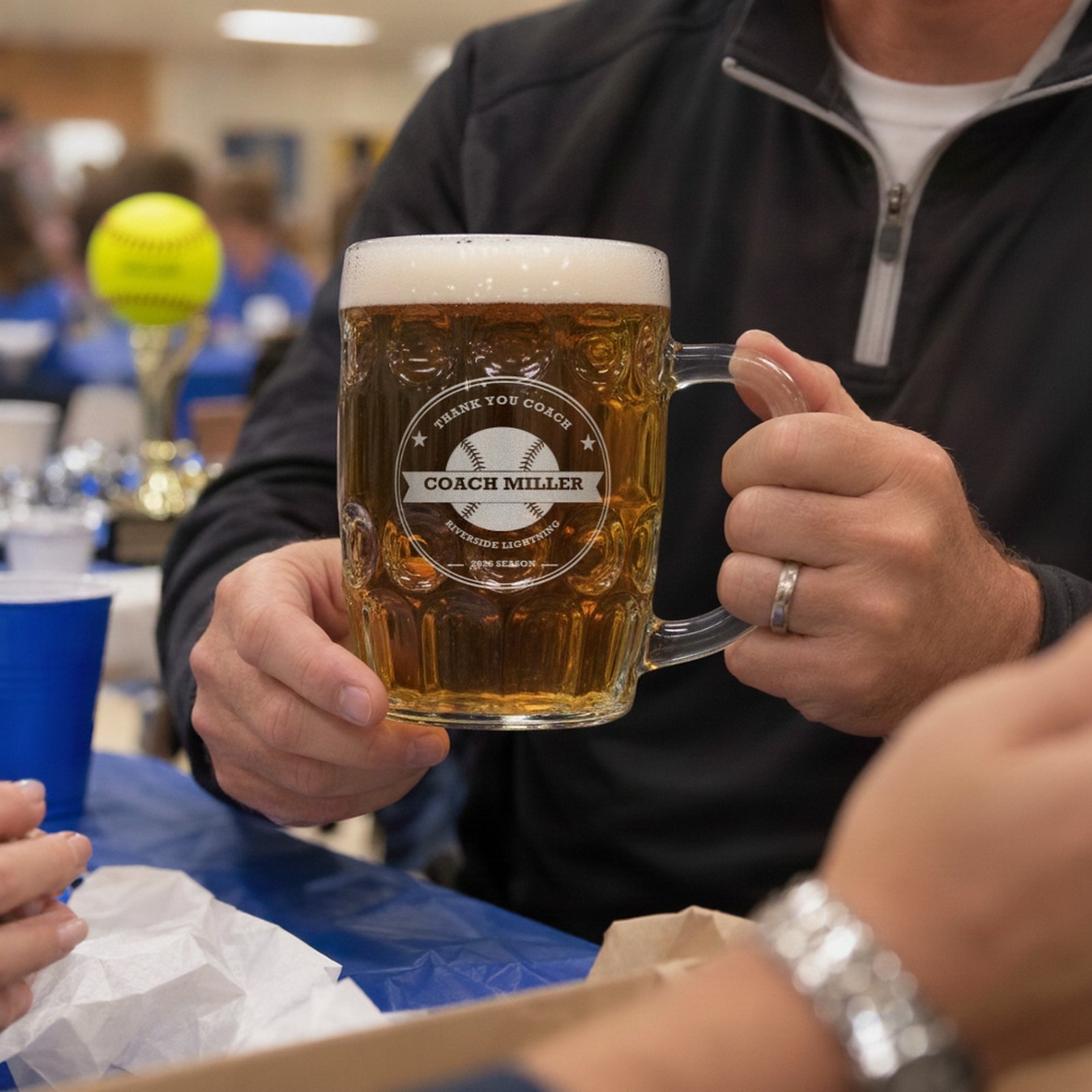 Coach holding the engraved softball coach beer stein at an end-of-season team banquet, a yellow softball trophy on the table in the background