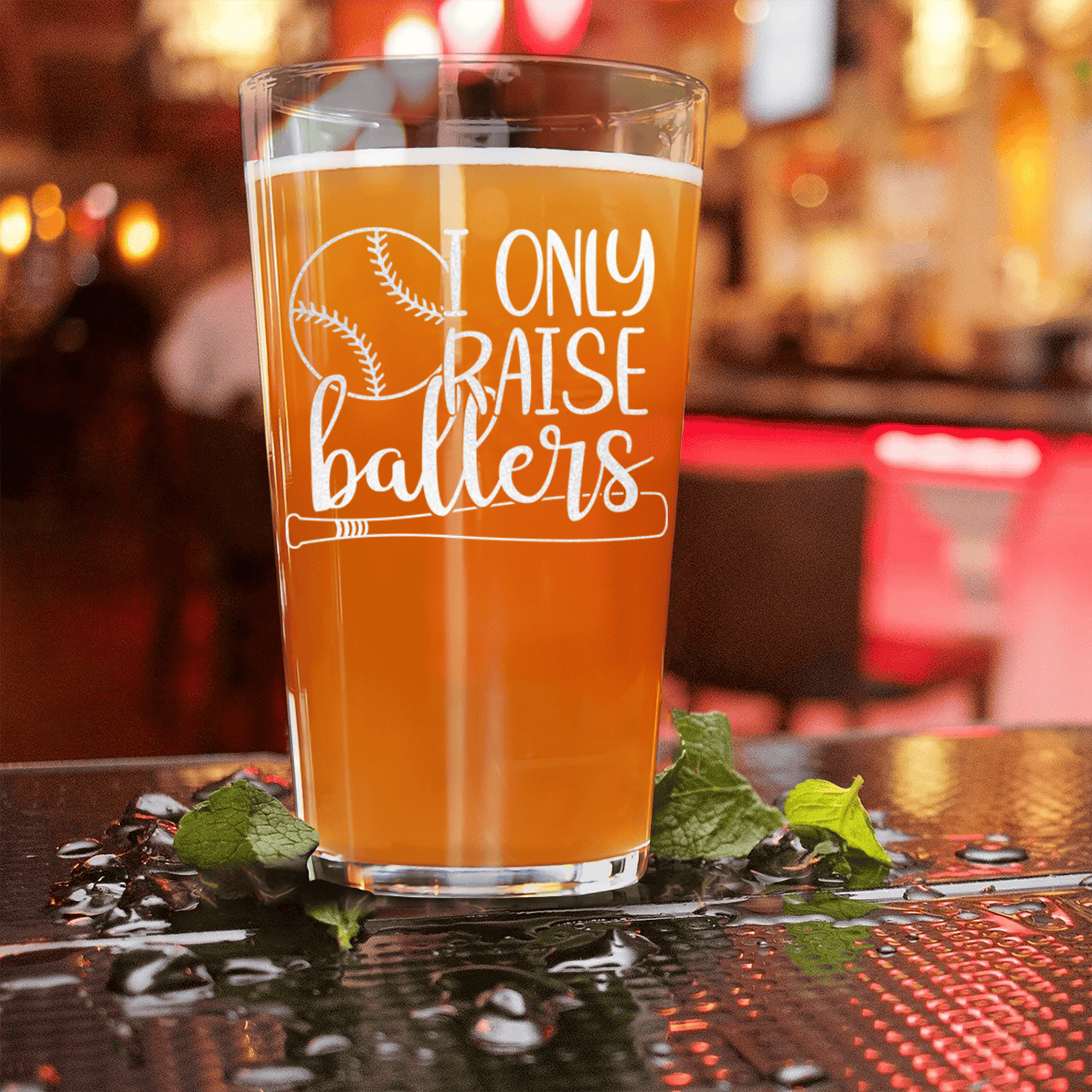 Pint glass filled with amber beer, etched with "I only raise ballers" text and baseball bat design, displayed on a bar counter with mint garnish and warm ambient lighting.