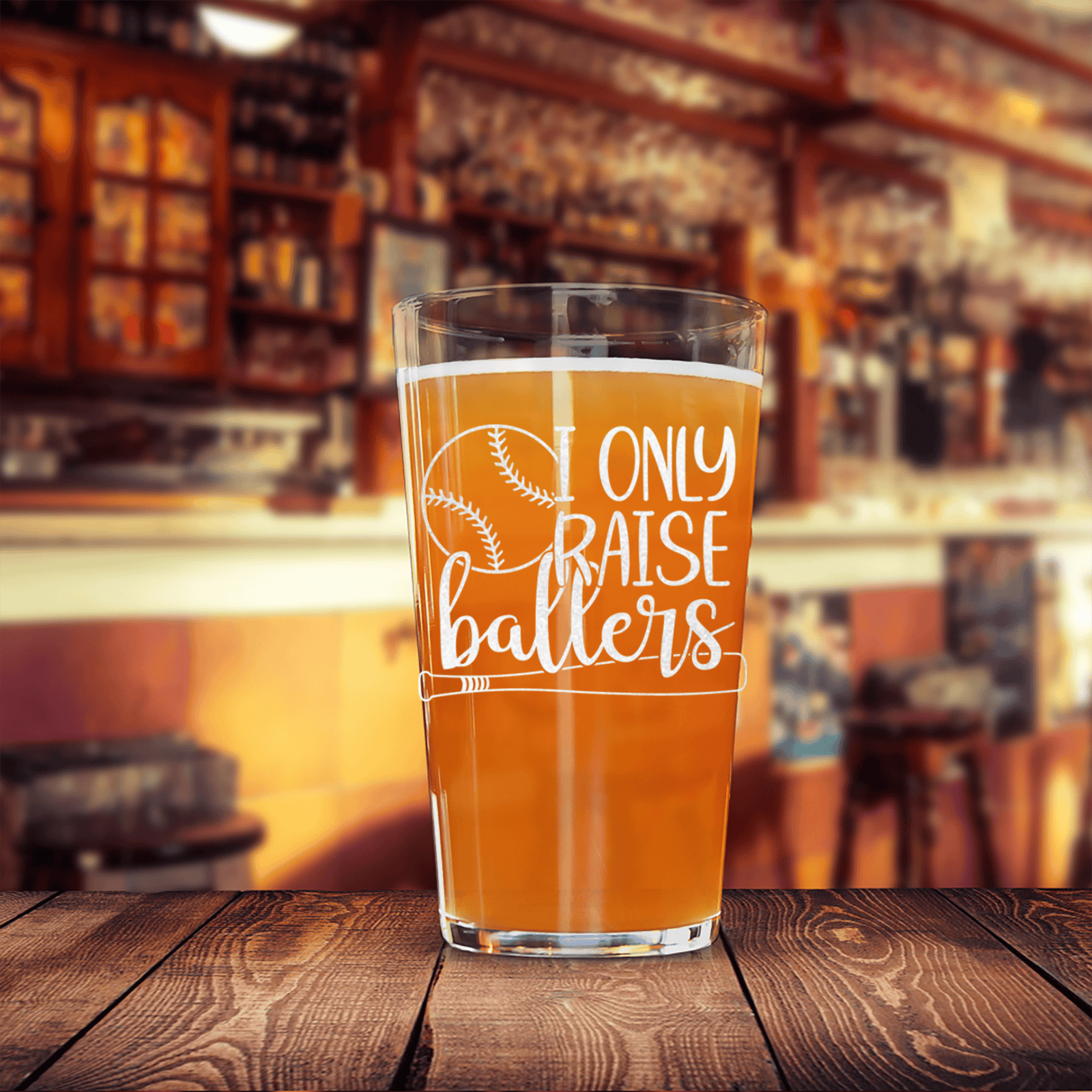 Clear pint glass etched with baseball and "I only raise ballers" text, filled with amber beverage, displayed on wooden surface with blurred bar background.