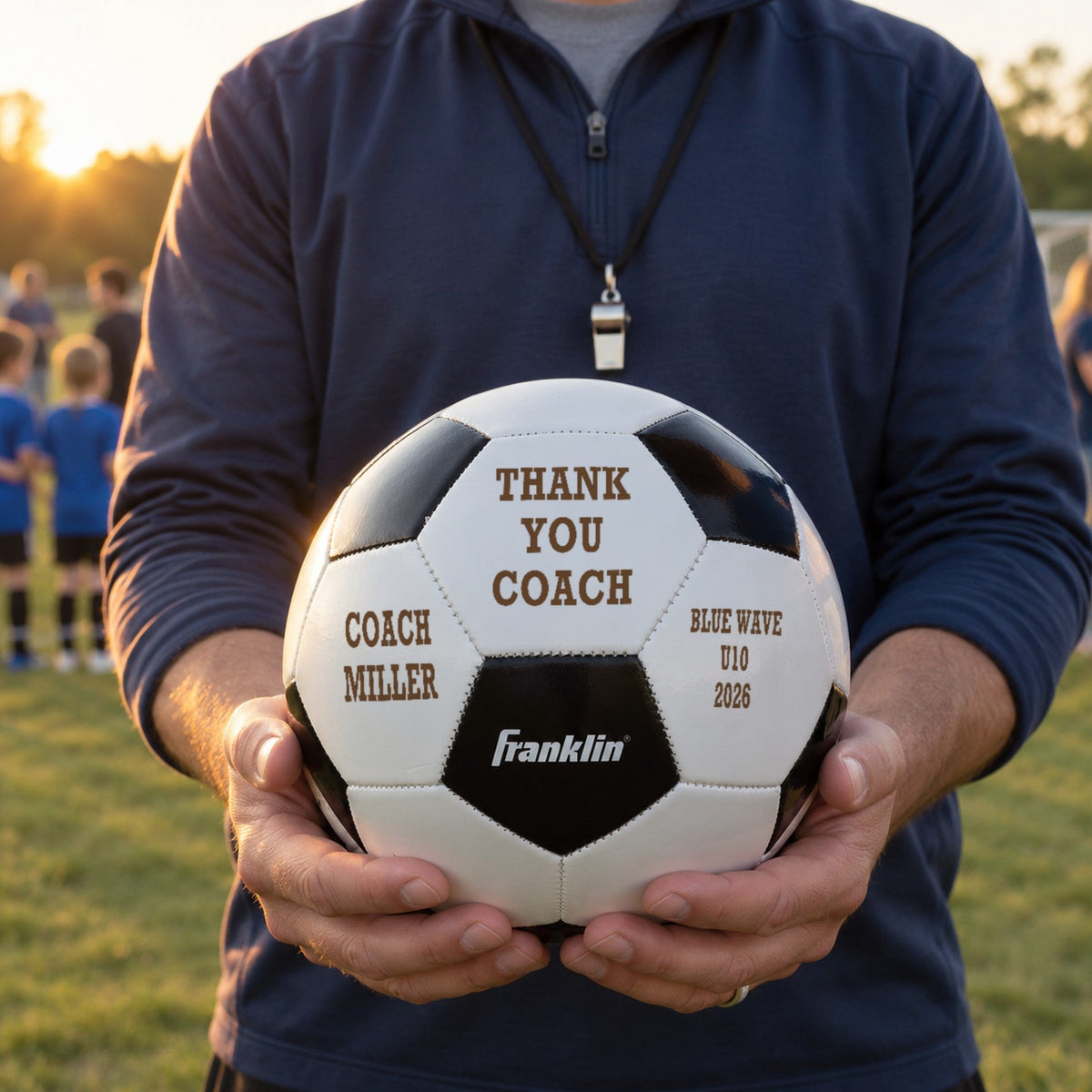 Youth soccer coach in a navy pullover holding an engraved ball that reads Thank You Coach, with the coach's name and team, after the last game