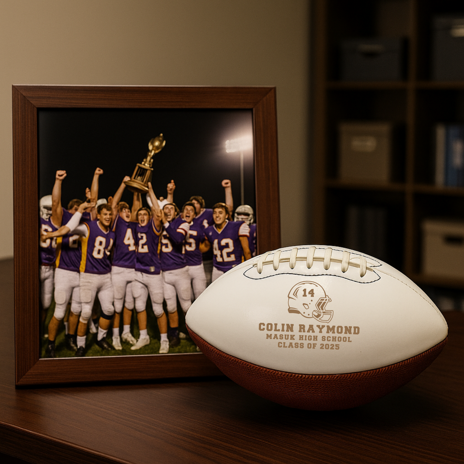 White leather football engraved with sample personalization showing name, number, school, and class year, displayed next to framed team photo