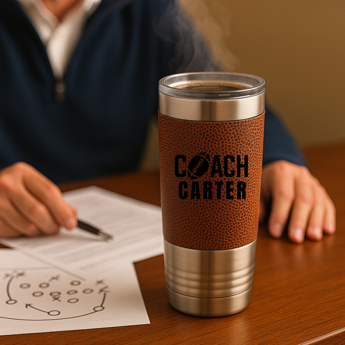 Tan leather-wrapped tumbler with stainless steel band engraved with sample text "COACH CARTER" and football icon, held by person at desk with football playbook sketch visible