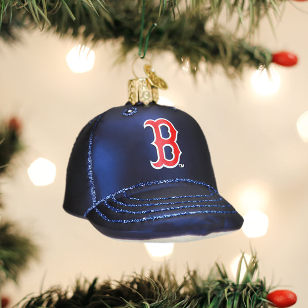 Navy baseball cap ornament with red Boston Red Sox "B" logo and glitter accent stripes, hanging from evergreen garland with lights
