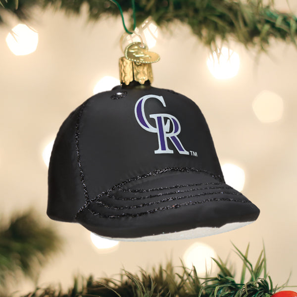 Black glass baseball cap ornament with purple and white Colorado Rockies logo, hanging from gold cap, surrounded by evergreen branches and white lights