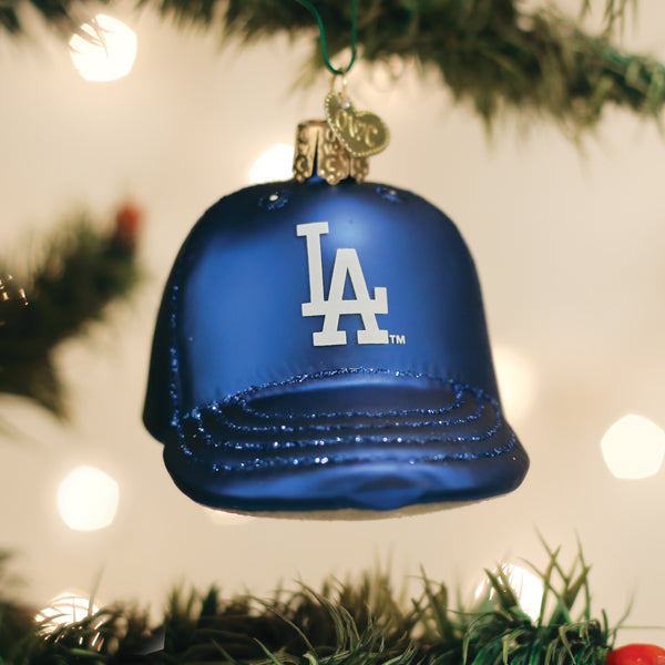 Blue baseball cap-shaped ornament with white LA logo, hanging from green ribbon against evergreen garland and bokeh lights