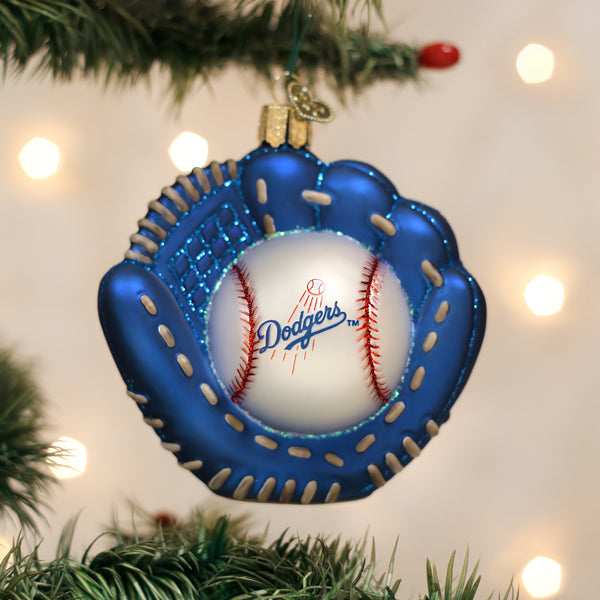 Blue baseball glove-shaped ornament with white baseball featuring Los Angeles Dodgers logo and red stitching, hanging from evergreen branch with warm lights in background
