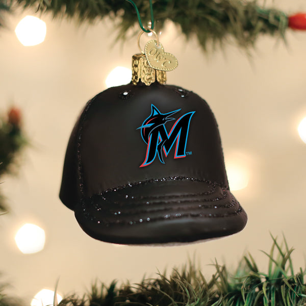 Black baseball cap ornament with teal and pink Marlins logo, embellished brim, hanging from green ribbon on evergreen branch with lights