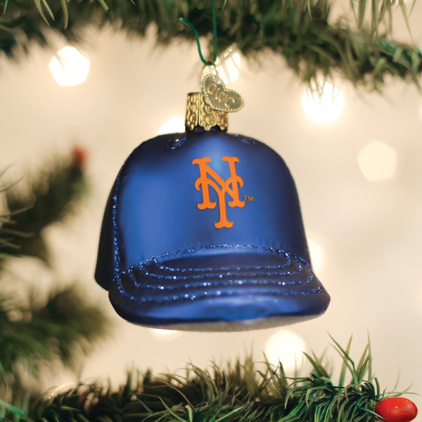 Blue baseball cap ornament with orange Mets logo, hanging from green ribbon on evergreen branch with white lights