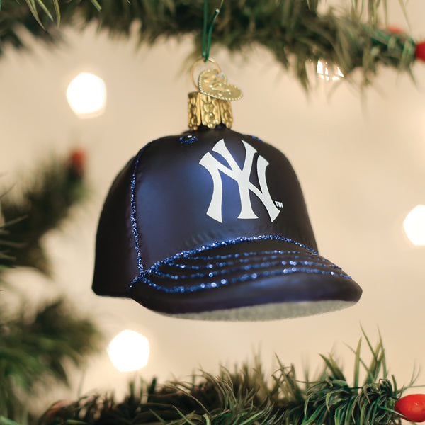 Navy blue baseball cap-shaped ornament with white "NY" logo and blue sequin bill, hanging from evergreen branch with blurred holiday lights in background