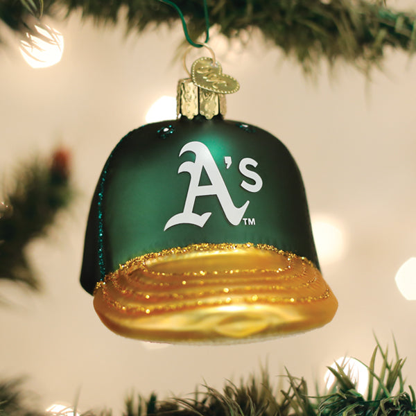 Green Oakland Athletics glass baseball-cap ornament with the white scripted 'A's' logo and gold glittered brim, hung on a lit Christmas tree