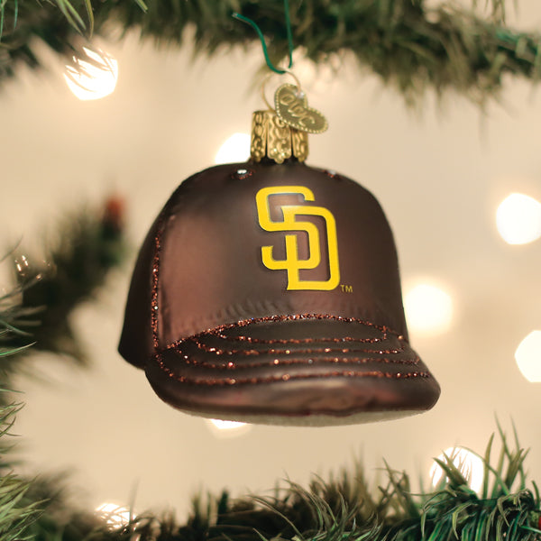 Brown and gold baseball cap ornament with Padres SD logo, hanging from green ribbon with evergreen branches and bokeh lights in background