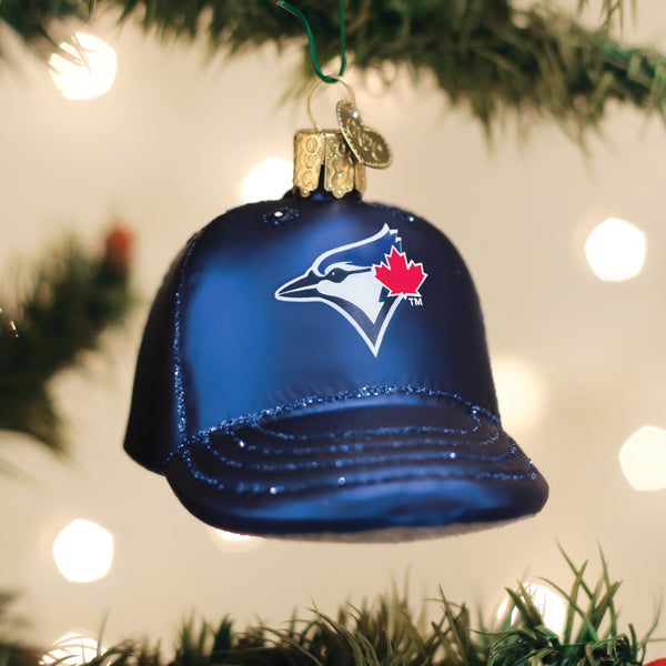 Blue glass baseball cap ornament shaped like a Toronto Blue Jays cap, featuring the team logo in white with red maple leaf, hanging from green ribbon among evergreen branches.