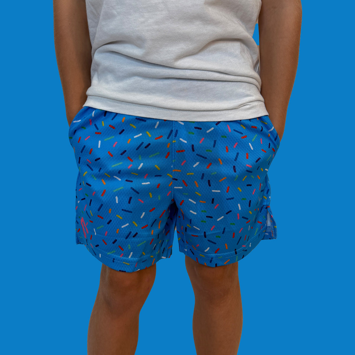 Man wearing blue shorts with multicolored sprinkle pattern and white t-shirt, shown from front waist down against blue background