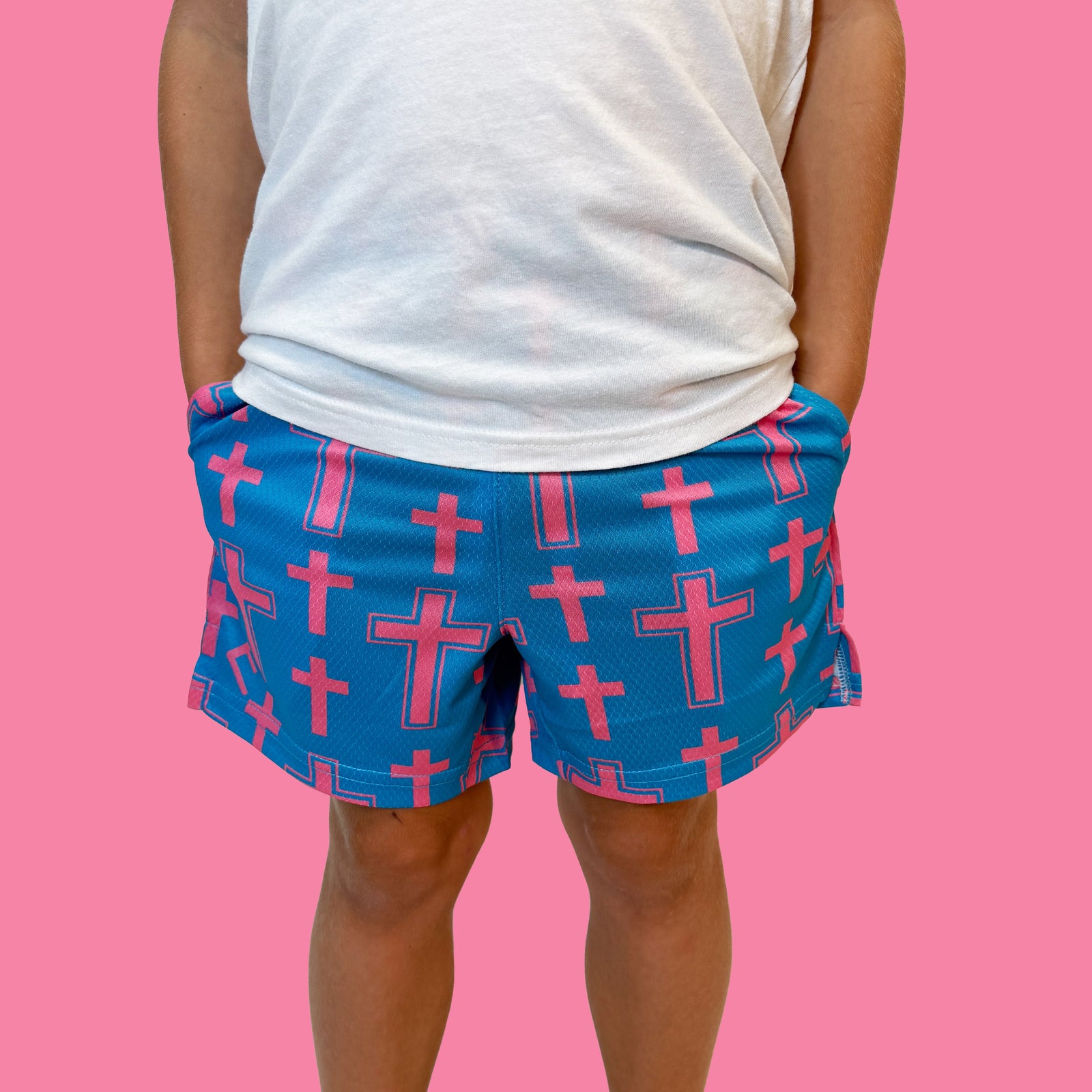 Person wearing white t-shirt and blue shorts with red cross pattern all over, shown from waist down against pink background