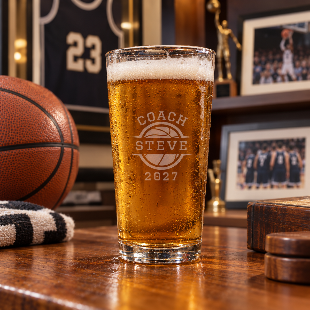 Clear pint glass filled with amber beer, engraved with sample text "Coach Steve" and "2027" inside a basketball circle, surrounded by sports decor