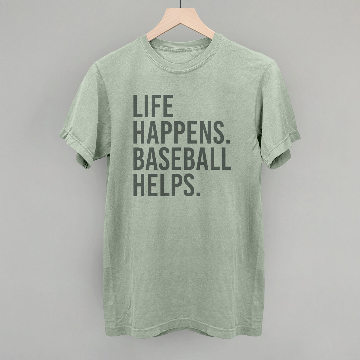 Sage green t-shirt with dark gray printed text reading "LIFE HAPPENS. BASEBALL HELPS." displayed on wooden hanger against gray background.
