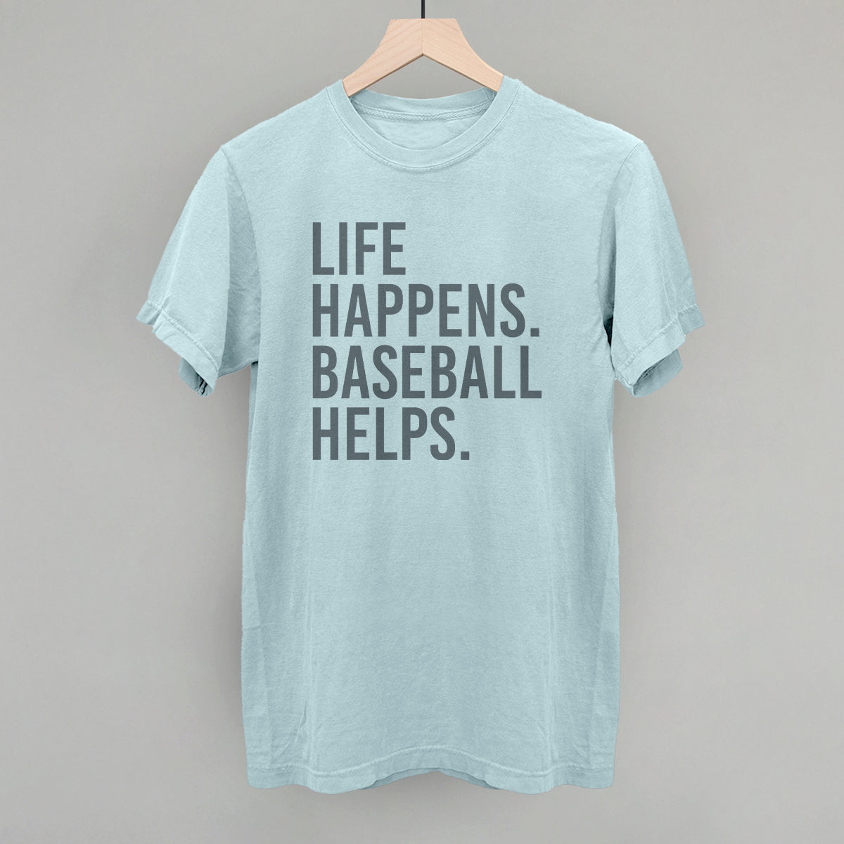 Light blue t-shirt with gray printed text reading "LIFE HAPPENS. BASEBALL HELPS." displayed on wooden hanger against neutral background