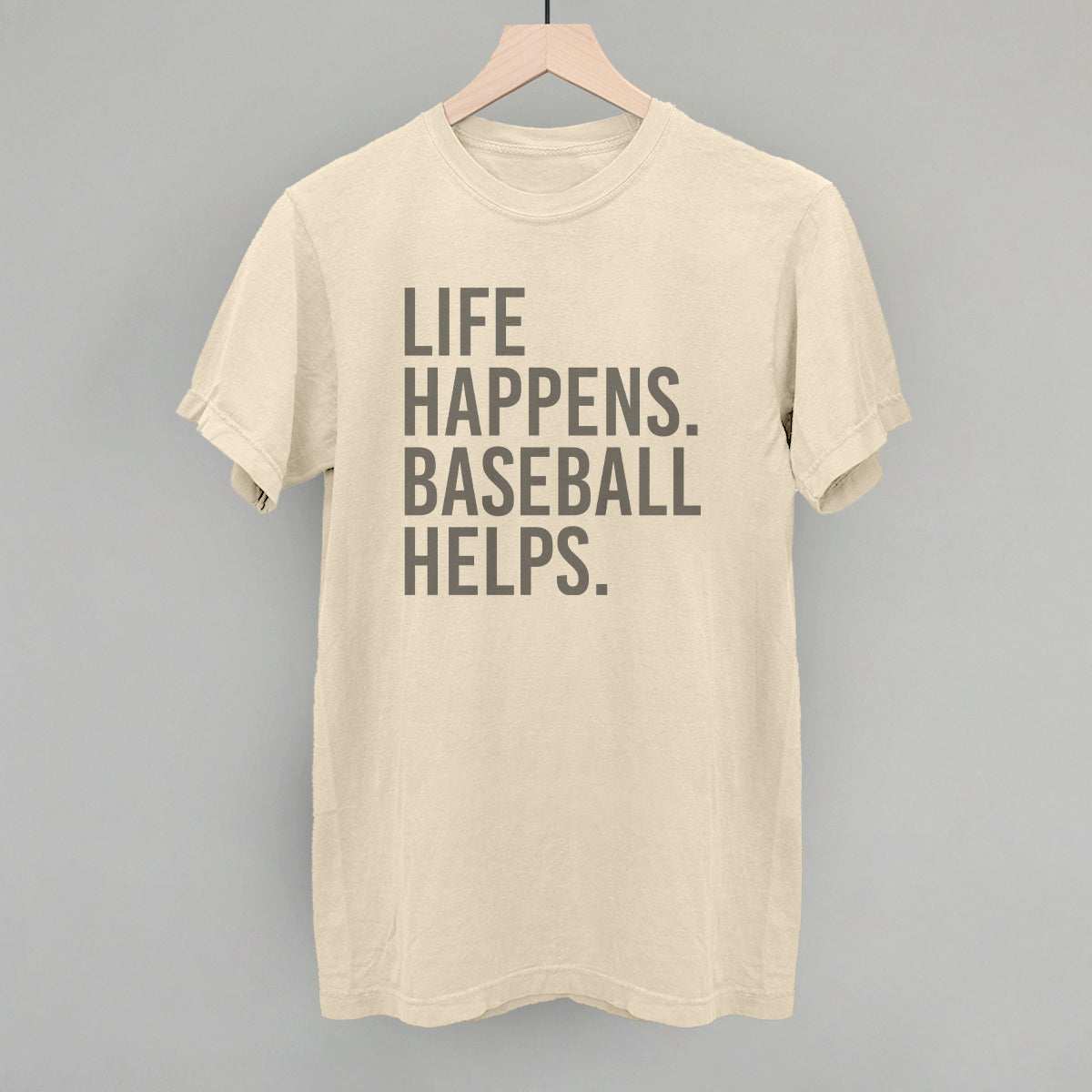 Cream-colored t-shirt with gray text reading "LIFE HAPPENS. BASEBALL HELPS." centered on the chest, displayed on a wooden hanger against a gray background.