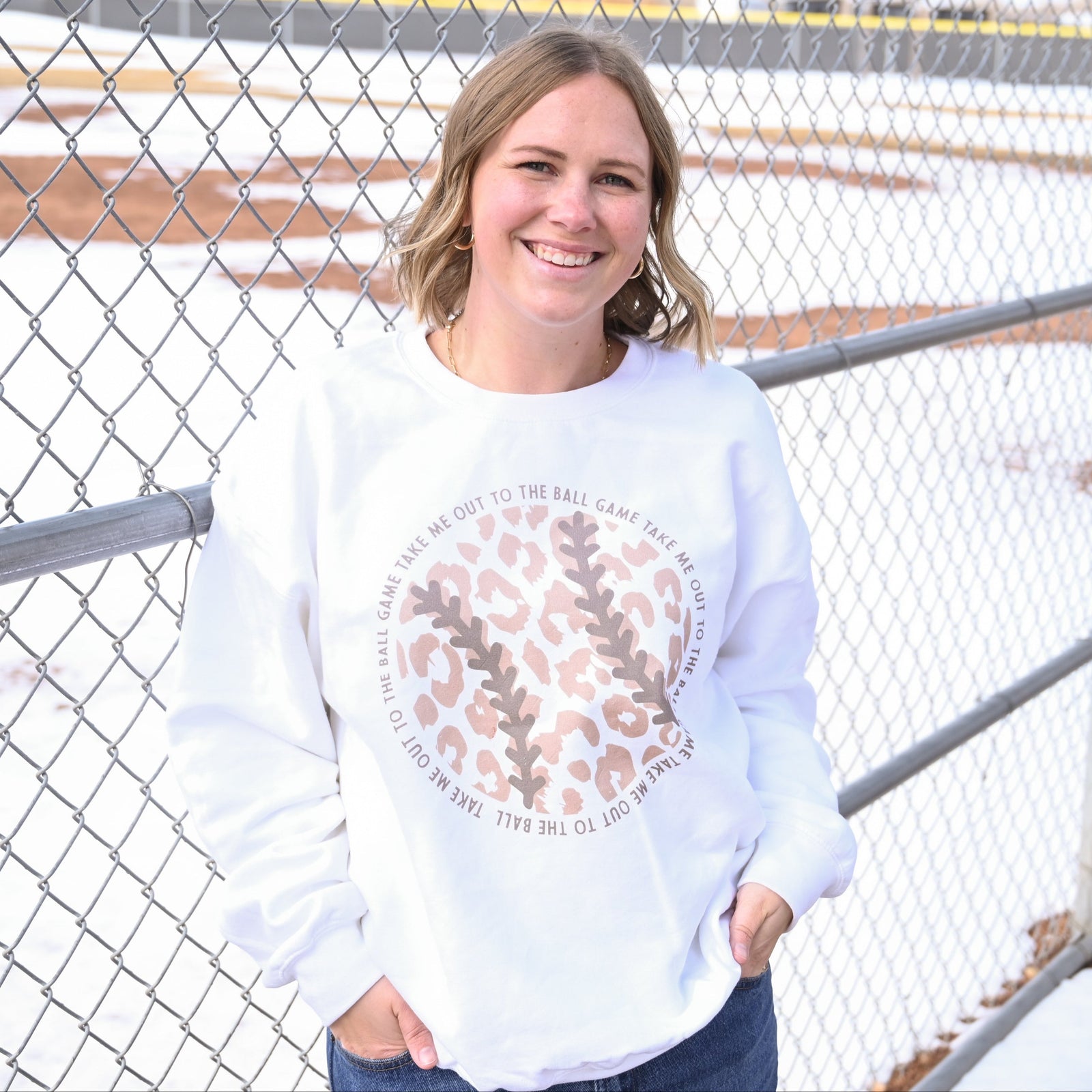 White crewneck sweatshirt with baseball leopard print and circular text reading "Take Me Out To The Ball Game" worn at a baseball field fence.