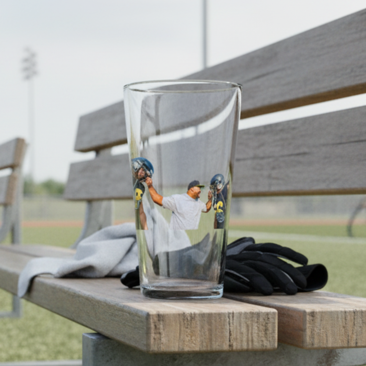 Pint glass printed with custom photo of baseball player mid-swing, displayed on wooden dugout bench with glove and baseball field in background